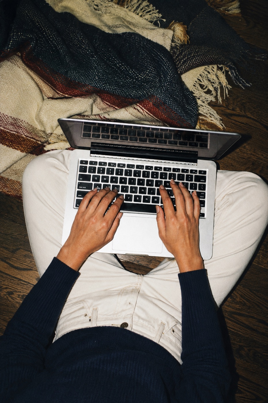 Someone cross-legged on the floor with a laptop, a blanket beside them, working late